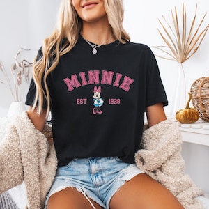 May include: Black t-shirt with the word "MINNIE" in pink block letters above a graphic of Minnie Mouse and the text "EST 1928". The shirt is worn with denim shorts and a cream-colored jacket.