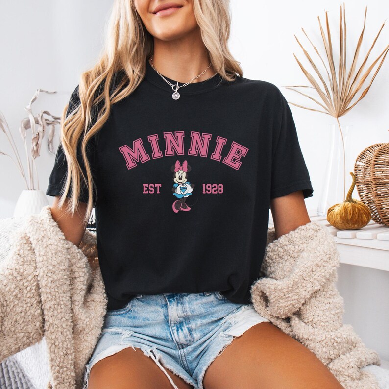 May include: Black t-shirt with the word "MINNIE" in pink block letters above a graphic of Minnie Mouse and the text "EST 1928". The shirt is worn with denim shorts and a cream-colored jacket.