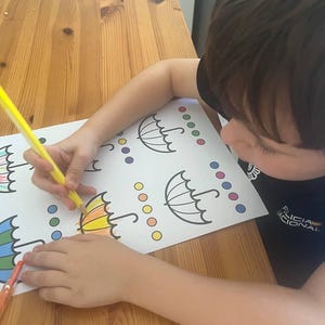 May include: A child coloring a printed page with umbrella outlines and colored circles. The child is using a yellow colored pencil to fill in the umbrella design. The table is made of wood.