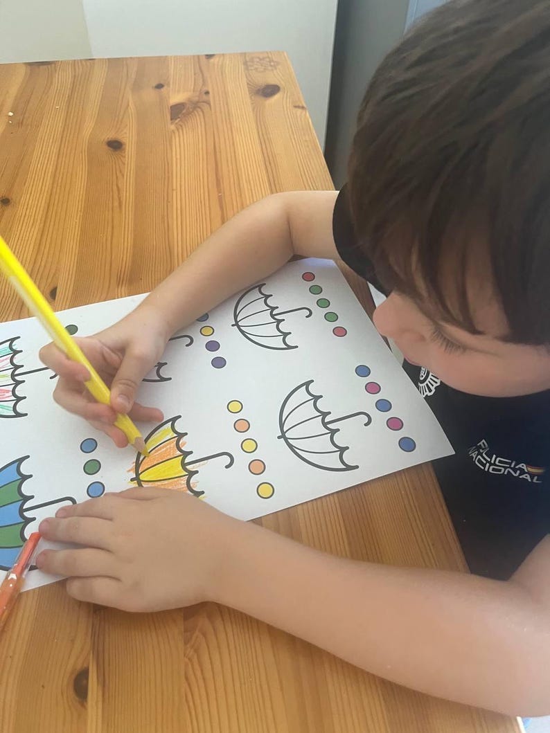 May include: A child coloring a printed page with umbrella outlines and colored circles. The child is using a yellow colored pencil to fill in the umbrella design. The table is made of wood.
