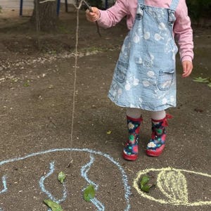 May include: A young child wearing a light blue denim dress with white floral patterns and colorful rain boots. The child is holding a stick with a string attached, standing near a chalk drawing on the ground.