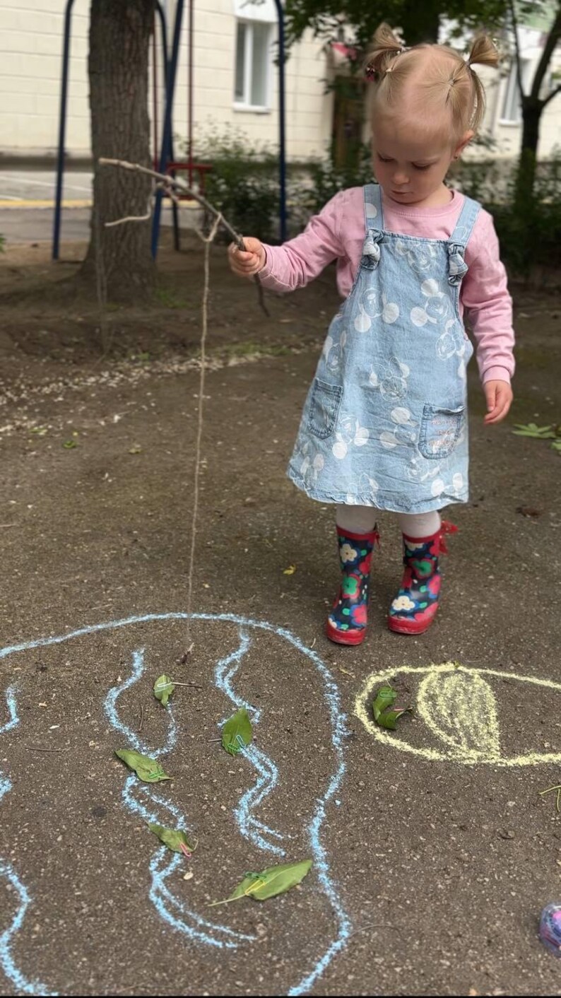 May include: A young child wearing a light blue denim dress with white floral patterns and colorful rain boots. The child is holding a stick with a string attached, standing near a chalk drawing on the ground.