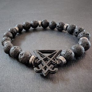 Peut inclure: Bracelet de perles en pierre de lave noire avec un charm géométrique noir et des entretoises argentées. Le bracelet présente des perles rondes et texturées et un charm central au design complexe. Un accessoire sombre et élégant.