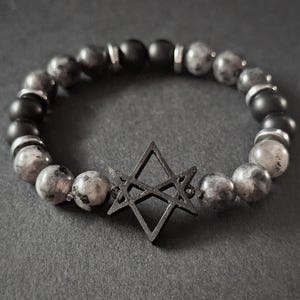 Può includere: Un braccialetto di perline con un ciondolo a forma di stella grigio scuro. Il braccialetto è composto da perline nere e grigie macchiate, con distanziatori color argento. Il braccialetto è esposto su una superficie grigio scuro.