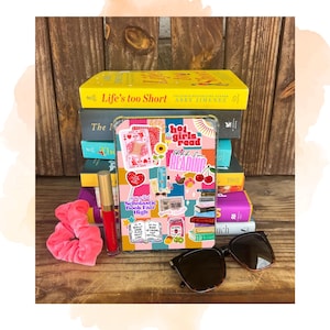May include: A stack of books with a decorative tablet case, sunglasses, a pink scrunchie, and a tube of red lip gloss. The tablet case has a colorful patchwork design with stickers that say "hot girls read" and "Scholastic Book Fair High".