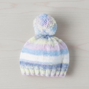 May include: A knitted beanie hat with a large pom-pom on top. The hat features horizontal stripes in pastel colors, including blue, white, lavender, and yellow. The pom-pom is a mix of light blue, lavender, and white. The hat is designed for warmth and style.