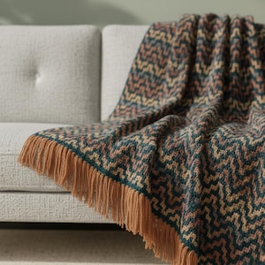 May include: A patterned throw blanket in teal, tan, and peach, with a geometric design, draped over a light grey sofa. The blanket features a tan fringe trim. The blanket is a cozy home decor item, perfect for adding warmth to a living space.