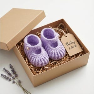 Può includere: Un paio di scarpine da neonato all'uncinetto color lavanda con un bottone a perla, presentate in una scatola regalo marrone con carta sminuzzata. Un'etichetta con le parole "Baby Love" è appesa alla scatola. Un rametto di lavanda è posto vicino alla scatola.