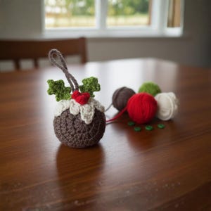 May include: A handmade crochet Christmas pudding ornament. The brown pudding is topped with white frosting, green holly leaves, and a red heart. Several balls of yarn in brown, red, green, and white are next to the ornament. Green beads are scattered on the table.