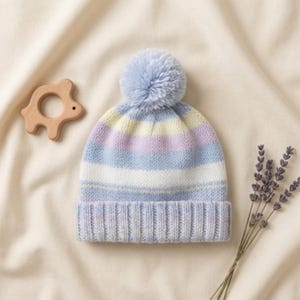 May include: A pastel-striped knit hat with a light blue pom-pom. The hat features horizontal stripes in shades of blue, white, yellow, and lavender. A wooden toy and sprig of lavender are also in the image.