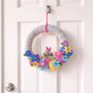 Puede incluir: Una corona de Pascua hecha a mano con una base envuelta en hilo pastel. La corona está adornada con coloridas flores de ganchillo, huevos de Pascua y una pequeña figura de conejito. La corona está colgada en una puerta blanca.