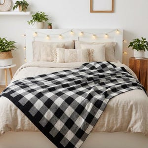 May include: A black and white checked blanket on a bed with beige bedding and pillows. A white headboard has fairy lights strung across it. Potted plants are on a shelf and a side table. The blanket's pattern is a large-scale check.