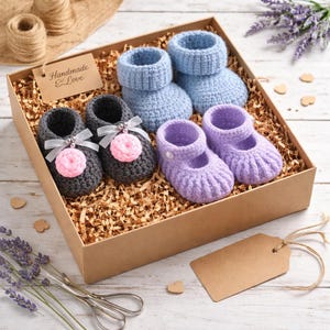 May include: A gift box containing three pairs of handmade crochet baby booties in gray, light blue, and lavender. The gray booties have pink pom-poms and bows. The box includes a tag that says "Handmade Love".
