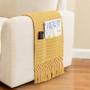 Puede incluir: Organizador de reposabrazos de ganchillo amarillo mostaza con un bolsillo para un mando a distancia y otro más grande que contiene una revista. La portada de la revista dice "COUNTRY FRENCH". El organizador tiene flecos en el borde inferior.