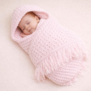 May include: A sleeping baby swaddled in a light pink crocheted blanket with a hood. The blanket features a fringe along the bottom. The baby is nestled inside, creating a cosy and warm environment.