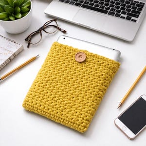 Puede incluir: Una funda de tableta de ganchillo amarillo mostaza con un cierre de botón de madera. La funda está hecha de hilo grueso y tiene un aspecto texturizado. Una tableta es parcialmente visible en el interior. Otros elementos en la superficie blanca incluyen un portátil, gafas y un smartphone.