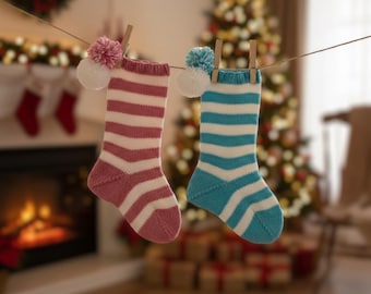 May include: Two knitted Christmas stockings, one pink and white striped, the other blue and white striped, hang from a clothesline. Each stocking has a pom-pom. A Christmas tree and fireplace are in the background.