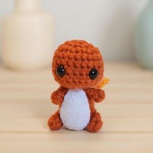 May include: A handmade orange crochet Charmander figure with black eyes and a white belly. The small amigurumi has a small yellow and orange flame on its tail. The toy is sitting on a light wooden surface.