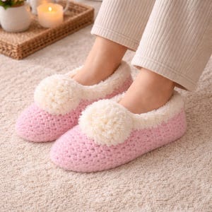 May include: Pink crochet slippers with cream-colored pom-poms and trim. The slippers have a soft, fuzzy interior. The image shows the slippers on a light-colored carpet.