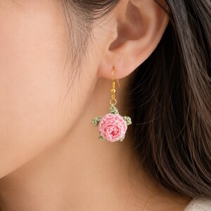 May include: A dangle earring featuring a small, crocheted pink rose with light green leaves. The floral element is attached to a gold-toned metal fishhook ear wire, creating a delicate accessory that hangs from the earlobe with a simple design.