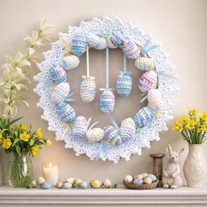 Puede incluir: Una corona de Pascua hecha a mano con huevos de ganchillo en colores pastel, adornada con cintas azules. La corona está rodeada por un tapete de encaje blanco. Decoraciones de Pascua adicionales incluyen un conejito, flores y huevos pintados.