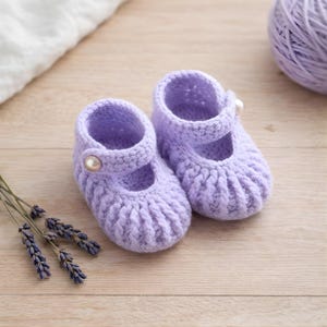 Puede incluir: Un par de zapatos de bebé de ganchillo lavanda hechos a mano. Los zapatos presentan un estilo Mary Jane con una correa y un cierre de botón perlado. Las suelas tienen una textura acanalada. Un ovillo de lana y ramitas de lavanda están en el fondo.