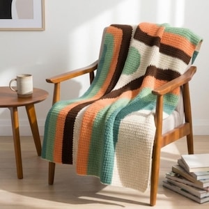 Puede incluir: Una manta de ganchillo a rayas en tonos naranja, marrón, verde y crema, sobre un sillón de madera. Una taza en una mesita auxiliar. La manta es un elemento de decoración del hogar.