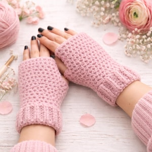 Op de afbeelding: Een paar lichtroze vingerloze handschoenen, gebreid met een textuurpatroon. De handschoenen hebben een geribbelde manchet en worden getoond op een licht oppervlak, met een bol wol en bloemen op de achtergrond.