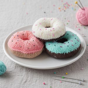 Op de afbeelding: Drie gebreide donuts op een wit bord. Eén is roze met kleurrijke hagelslag, een andere is wit met roze hagelslag, en de derde is turquoise met hagelslag. Allemaal hebben ze bruine bodems en zijn handgemaakt.