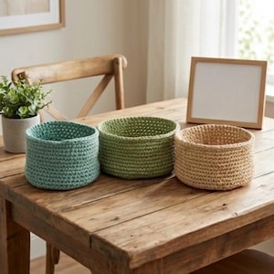 May include: Three handmade crochet baskets in shades of blue, green, and tan sit on a wooden table. The baskets are cylindrical with a textured stitch pattern. A small potted plant and a framed blank picture are also on the table.