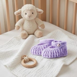 May include: A cream-coloured lamb toy sits on a white blanket next to a pair of lavender crochet baby booties. A wooden teething ring rests on the blanket. The items are arranged on a cot.
