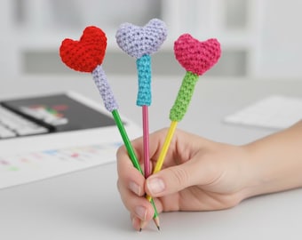 May include: Three pencils with crochet heart toppers in red, purple, and pink. The pencils have green, pink, and yellow shafts. The crochet toppers have coordinating colors. The pencils are held in a hand, with a desk and keyboard in the background.