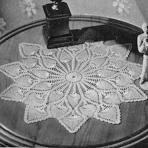 May include: An off-white, star-shaped crochet doily with an intricate design. The doily is placed on a dark wooden table, with a decorative figurine and a lamp base in the background. The doily's pattern features a central circle and radiating designs.
