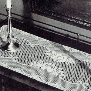 May include: A white crocheted table runner with a delicate floral design. Two silver candle holders with candles are placed on the runner.