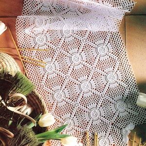 Può includere: Un runner da tavolo bianco all'uncinetto con un delicato motivo floreale. Il runner è realizzato con un filo sottile e ha un aspetto traforato. È disposto su una superficie di piastrelle marroni con un cesto di fiori e verde in primo piano.