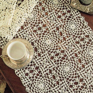 May include: A white crocheted table runner with a delicate floral pattern. The runner is laid on a wooden table with a silver teacup and saucer and a framed photograph.