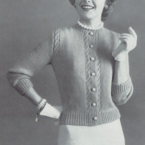 Op de afbeelding: Een vrouw draagt een lichtbruine gebreide cardigan met een kabelbreipatroon en parelknopen. Ze draagt een witte rok en een parelketting.