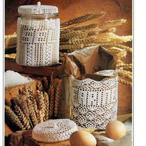 May include: A still-life image featuring a glass jar and a container, both adorned with intricate white crochet. The jar has a lid, and the container has the word "BREAD" crocheted on it. Wheat stalks and eggs are also present.