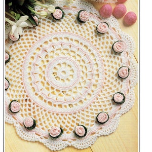 Può includere: Tovaglietta all'uncinetto bianca con un centro rosa e dettagli di fiori rosa attorno al bordo. La tovaglietta è su una superficie di legno.