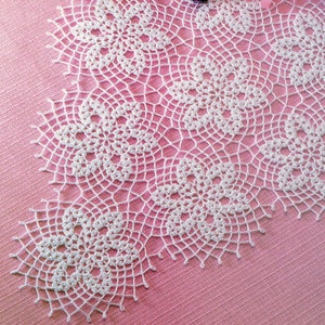 May include: White crocheted doily with a delicate floral pattern on a pink background. The doily is made of fine thread and has a scalloped edge.