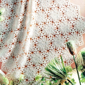 May include: A white crocheted doily with a delicate floral pattern. The doily is laid out on a pink surface with green plants and a wicker basket.