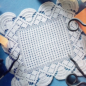 May include: White crocheted doily with a square center and scalloped edges. The doily is laid on a blue surface with a black metal object.