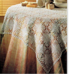 May include: A white, openwork crochet tablecloth with a floral pattern covers a round table. The tablecloth is draped over a table with a patterned fabric. The words "Vintage Stitchery Archives" are on the left side.