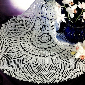 Puede incluir: Un mantel de mesa de crochet blanco con un delicado patrón floral. El mantel está drapeado sobre un jarrón azul con orquídeas blancas.