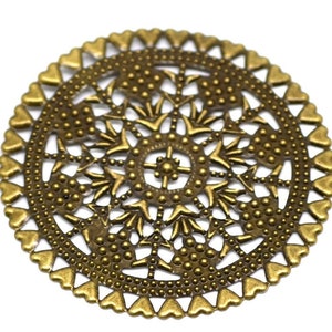 May include: A round, gold-colored metal charm with a detailed, intricate design featuring hearts, dots, and swirls. The charm has a filigree pattern and a slightly raised center.