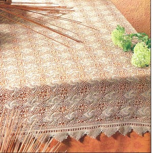 May include: Cream-colored, intricately crocheted tablecloth with a detailed floral pattern. The tablecloth has a scalloped edge and is adorned with decorative elements. Dried stems and green flowers are placed on the table.