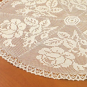Könnte beinhalten: Cremefarbene, runde gehäkelte Tischdecke mit floralem Muster. Das filigrane Design zeigt sich wiederholende Blumen- und geometrische Formen mit einem gewellten Rand. Die Tischdecke besteht aus einem zarten, offen gewebten Stoff.