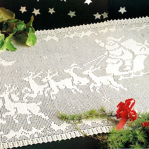 Può includere: Un runner da tavolo in crochet bianco con un design di Babbo Natale e renne che tirano una slitta. Il runner è circondato da rami di pino verdi e fiocchi rossi.