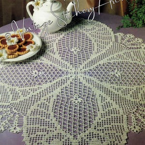 May include: A white crocheted table runner with a delicate floral design. The runner is laid out on a purple table with a teapot and cookies on a plate.