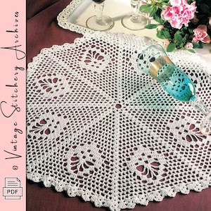 May include: A round white crochet doily featuring a repeating fan-like pattern and a scalloped edge. The doily is displayed on a dark surface with a teal glass flute and a white tray with two clear glasses and pink flowers nearby.